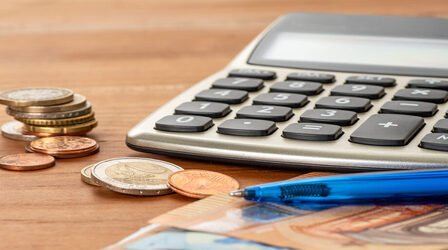 Man sieht einen Taschenrechner, davor liegen Münzen und ein Kugelschreiber