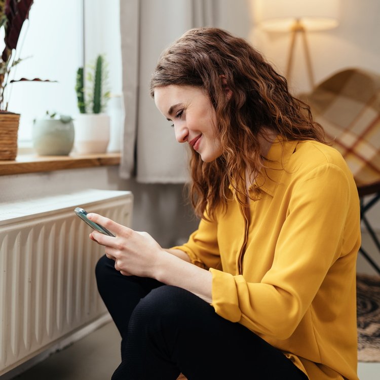 Frau am Smartphone vor einer Heizung sitzend.