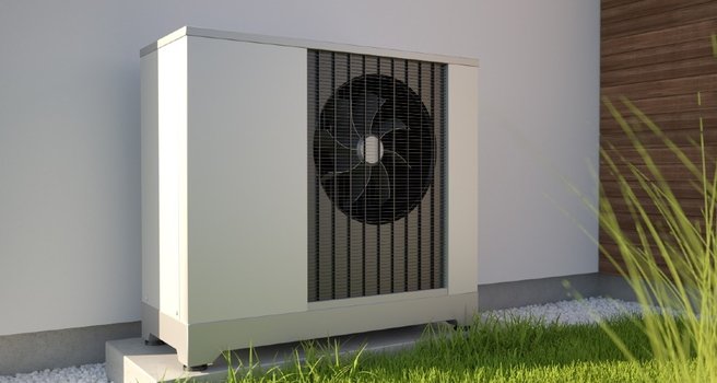 Die Außeneinheit einer Wärmepumpe ist vor einer weißen Außenfassade im Garten eines Einfamilienhauses zu sehen.