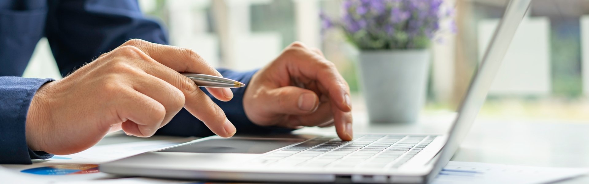Mann sitzt vor einem Laptop mit einem Kugelschreiber in der Hand wie er eine Eingabe auf der Tastatur macht