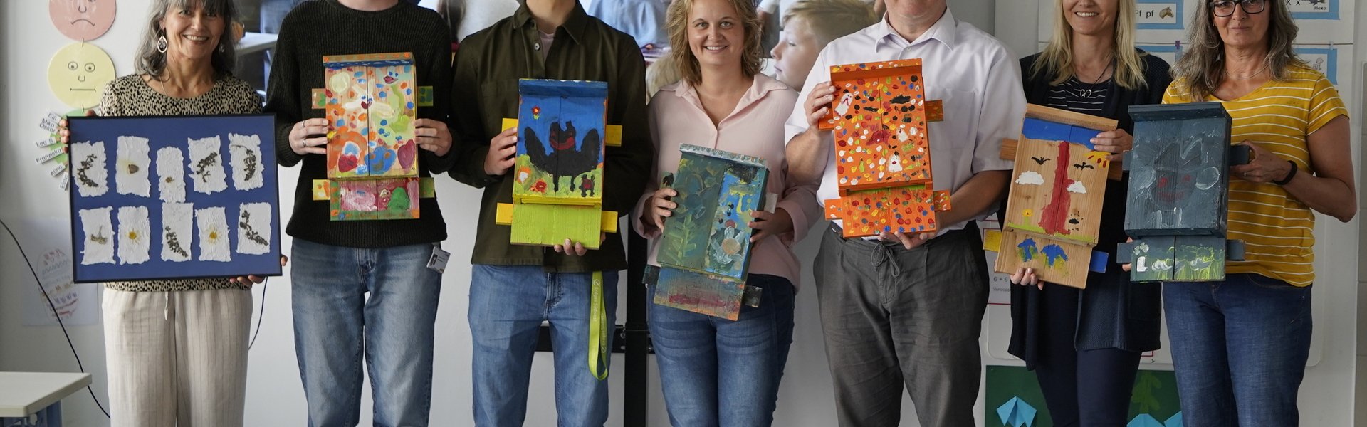 Personengruppe in einem Klassenzimmer hält selbst gestaltete Kunstwerke vor einer Präsentation hoch.