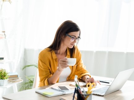 Eine Frau sitzt in einem hellen Büro an einem Schreibtisch vor ihrem Laptop, Notizen und Büromaterial, in der Hand eine Tasse Kaffee.