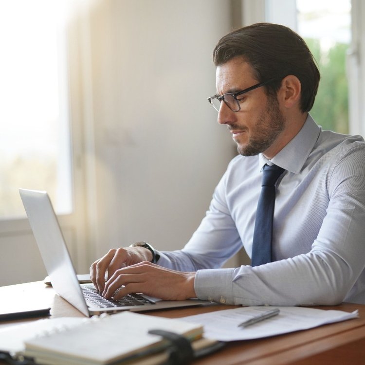 Ein Geschäftsmann im Anzug sitzt am Schreibtisch vor seinem Laptop und gibt über seine Tastatur etwas ein