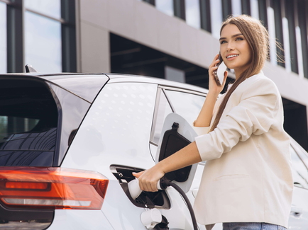 Eine junge Frau telefoniert am Handy, während sie ihr Elektrofahrzeug lädt.