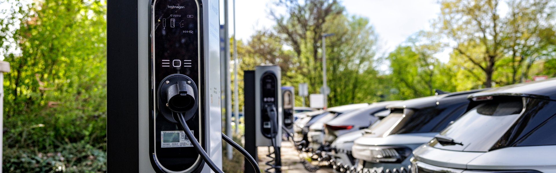 Elektroauto lädt an einer Ladesäule auf einem Parkplatz mit mehreren geparkten Fahrzeugen im Hintergrund.