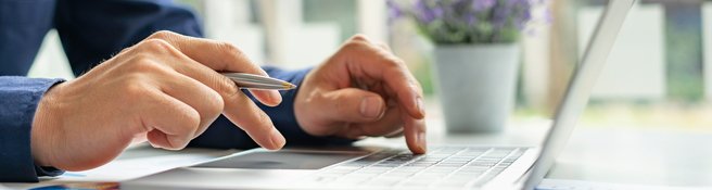 Mann sitzt vor einem Laptop mit einem Kugelschreiber in der Hand wie er eine Eingabe auf der Tastatur macht