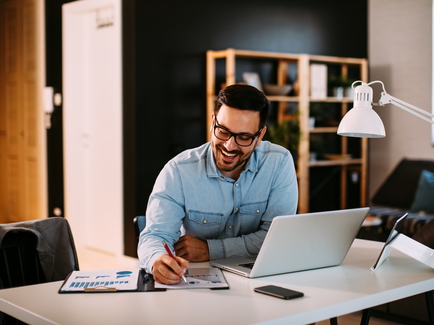 Mann mittleren Alters sitzt an einem Schreibtisch vor einem Laptop, Unterlagen und einem Smartphone und macht mit einem Kugelschreiber eine Markierung in seinen Unterlagen.