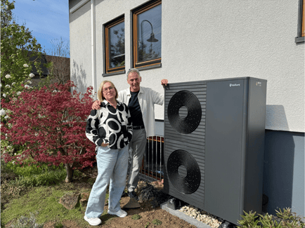 Ehepaar steht neben einer modernen Vaillant-Luft-Wasser-Wärmepumpe vor einem Wohnhaus mit Garten