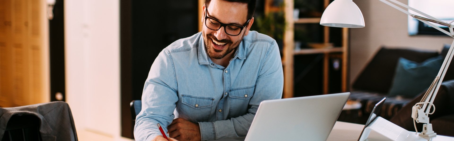 Mann mittleren Alters sitzt an einem Schreibtisch vor einem Laptop, Unterlagen und einem Smartphone und macht mit einem Kugelschreiber eine Markierung in seinen Unterlagen.